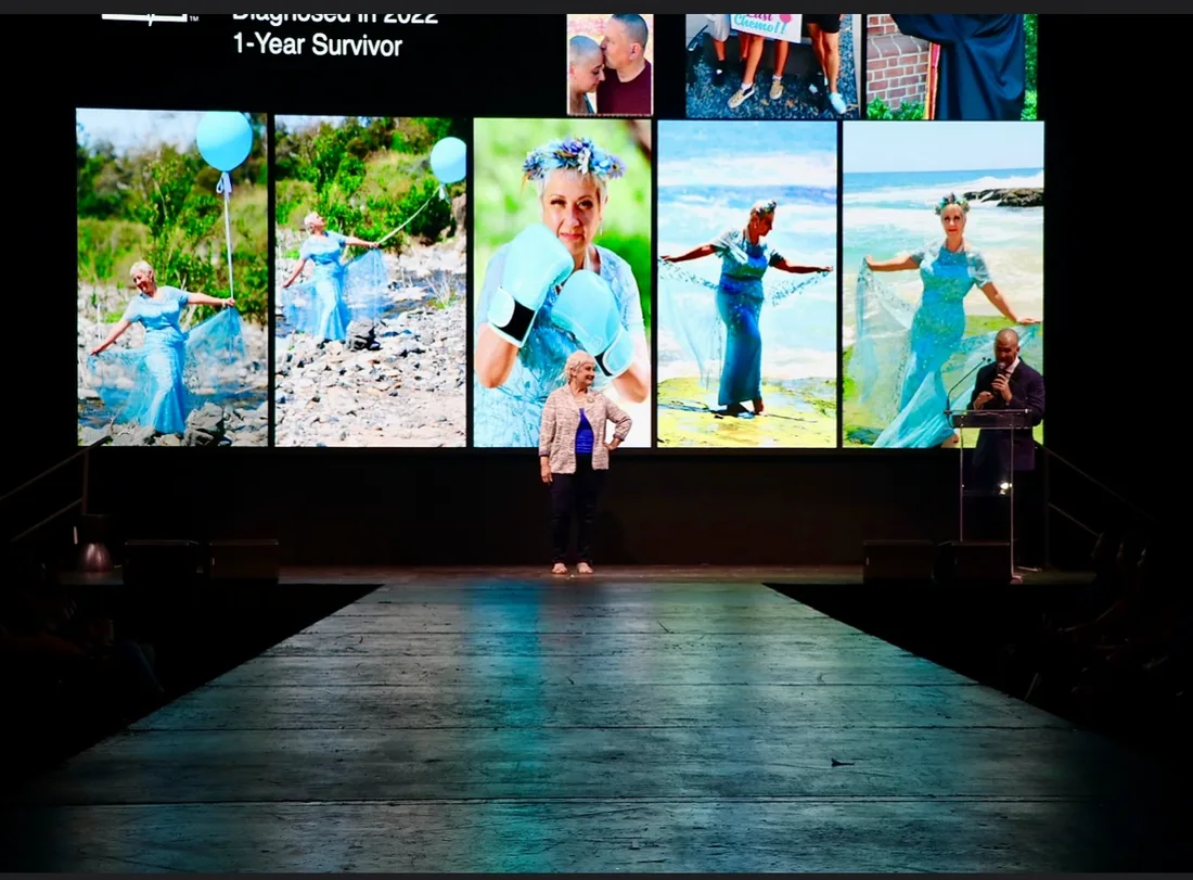 Tina standing at the end of the runway at a survivor fashion show, photographs from her cancer journey filling the screens behind her.